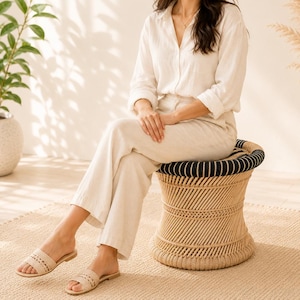May include: A woven rattan stool with a black and white striped top. The stool has a rounded shape and is made of natural-coloured rattan. The stool is approximately 46 cm tall and 41 cm in diameter.