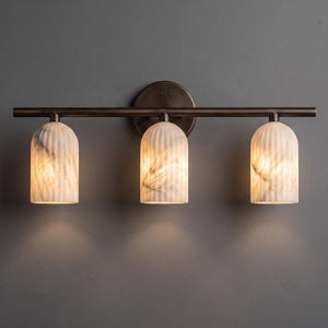 May include: A wall-mounted light fixture featuring a horizontal dark bronze metal bar and a circular backplate. Three cylindrical, vertically ribbed glass shades with a marbled white and grey pattern are attached, each emitting a warm glow.