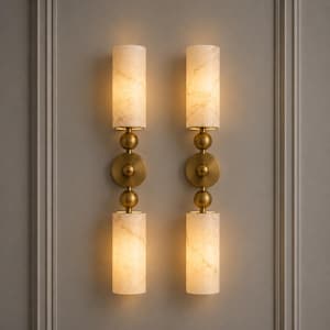May include: A pair of wall sconces featuring two cylindrical, cream-colored stone shades each, separated by a central brass circular mount and two brass spheres. The fixtures emit a warm, soft glow from the top and bottom stone cylinders.