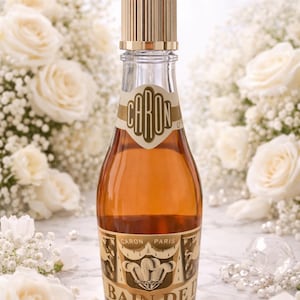 May include: A clear glass bottle of Caron Bain de Je perfume with a gold-coloured cap. The bottle has a gold and beige label with the brand name and product name. The background features white roses and baby's breath.