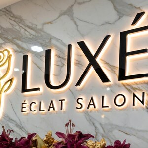 May include: Illuminated sign for LUX&Eacute; &Eacute;CLAT SALON, featuring a gold rose and black letters with a warm glow against a marble background. The salon's name is displayed prominently, with the word LUX&Eacute; above &Eacute;CLAT SALON.
