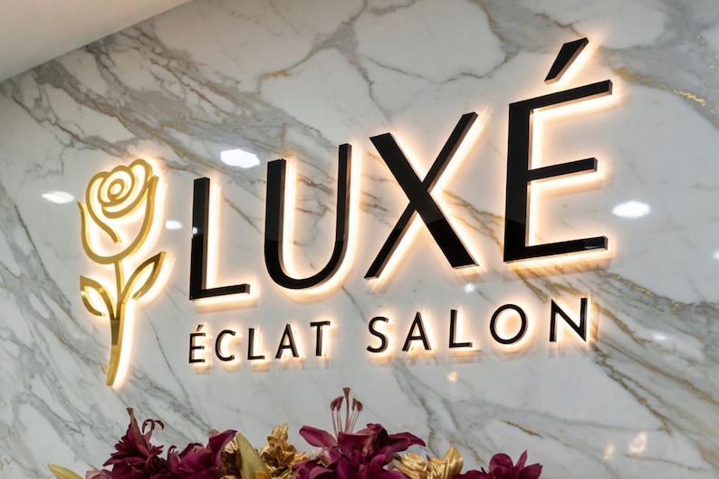May include: Illuminated sign for LUX&Eacute; &Eacute;CLAT SALON, featuring a gold rose and black letters with a warm glow against a marble background. The salon's name is displayed prominently, with the word LUX&Eacute; above &Eacute;CLAT SALON.