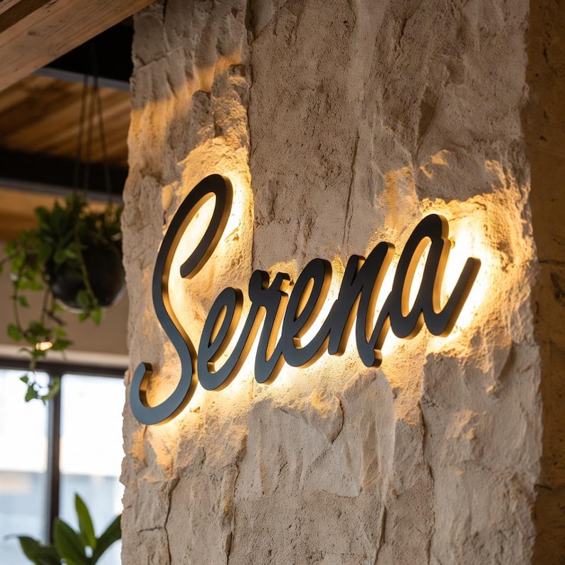 May include: A black, cursive-style metal sign displaying the name "Serena" mounted on a textured, light-colored stone wall. The sign features integrated backlighting that casts a warm, glowing halo effect around the letters against the stone.