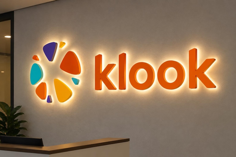 May include: A wall-mounted sign featuring the word klook in orange 3D letters alongside a circular logo composed of various orange, yellow, teal, and purple geometric shapes. The entire display is backlit, casting a soft glow against the wall.