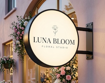 May include: A circular, illuminated storefront sign with a black frame mounted on a wall. The sign features a black line-art flower icon above the text LUNA BLOOM and the words FLORAL STUDIO in a clean, black, sans-serif typeface.