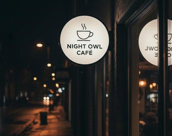May include: A circular, illuminated sign mounted on a brick wall displays the text NIGHT OWL CAFÉ in black lettering beneath a simple line drawing of a steaming coffee cup. The sign glows against a dark, nighttime street background.
