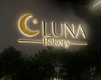 May include: An illuminated wall sign for LUNA Bakery featuring a gold crescent moon and a four-pointed star icon to the left of the word LUNA in large capital letters, with the word Bakery centered below, all backlit with a warm glow.