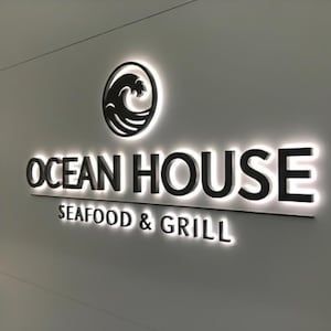 May include: Illuminated sign for "Ocean House Seafood & Grill" on a light-colored wall. The sign features black lettering and a wave logo within a circle. The words "Seafood & Grill" are below the main text.
