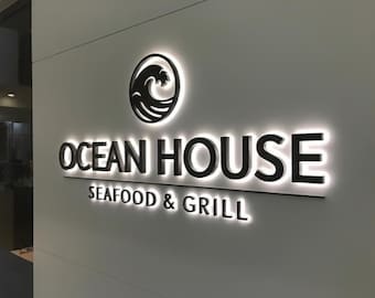 May include: Illuminated sign for "Ocean House Seafood & Grill" on a light-colored wall. The sign features black lettering and a wave logo within a circle. The words "Seafood & Grill" are below the main text.