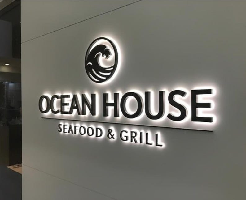 May include: Illuminated sign for "Ocean House Seafood & Grill" on a light-colored wall. The sign features black lettering and a wave logo within a circle. The words "Seafood & Grill" are below the main text.