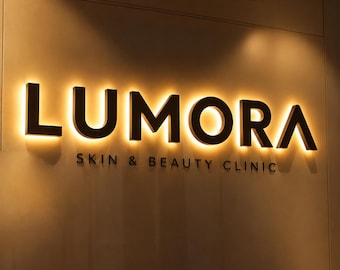 May include: A wall-mounted sign featuring the word LUMORA in large, dark, three-dimensional letters with a warm, golden backlighting effect. Below the main text, the words SKIN & BEAUTY CLINIC are displayed in smaller, dark, flat lettering.