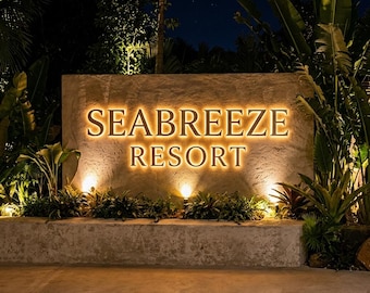 May include: A rectangular stone sign with the text SEABREEZE RESORT illuminated in warm yellow light. The sign is set against a dark background with lush green tropical plants and small ground-level spotlights at the base.