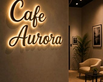May include: A wall-mounted sign with the text Cafe Aurora in a dark, cursive font. The letters are backlit with a warm, glowing light that casts a soft halo effect against the textured, light-colored curved wall surface.