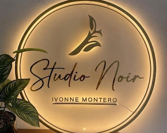 May include: A circular, backlit wall sign with a gold frame. The sign features a stylized leaf icon at the top, the text "Studio Noir" in a cursive script, and the text "IVONNE MONTERO" in a clean, sans-serif font with a horizontal line below.