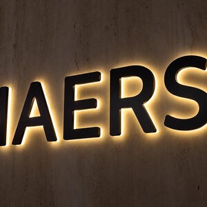 May include: A wall-mounted sign featuring the word NAERSI in bold, black, sans-serif capital letters. Each letter is backlit with a warm, glowing light that creates a soft halo effect against the textured, neutral-toned wall surface behind it.