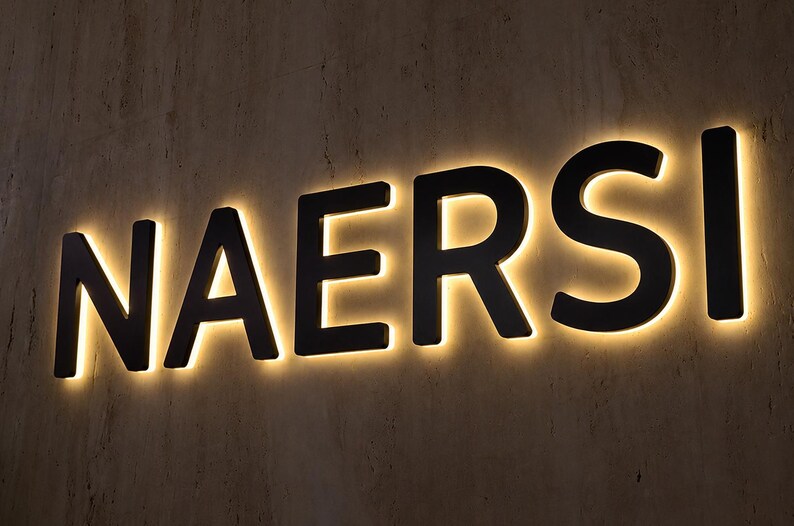 May include: A wall-mounted sign featuring the word NAERSI in bold, black, sans-serif capital letters. Each letter is backlit with a warm, glowing light that creates a soft halo effect against the textured, neutral-toned wall surface behind it.