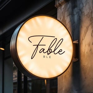 Puede incluir: Un letrero circular de pared iluminado con una cara blanca que emite un brillo cálido. La palabra Fable está escrita en una fuente cursiva negra en el centro, con las letras BLE impresas debajo en una fuente sans-serif más pequeña.