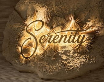 May include: A wall-mounted sign featuring the word Serenity in a cursive script, illuminated by warm backlighting. The sign is set against a textured, off-white, irregular stone-like base adorned with numerous dried, fan-shaped palm leaves.