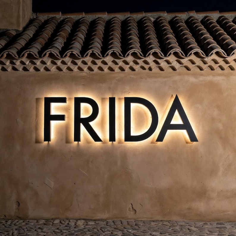 May include: Illuminated sign spelling "FRIDA" in black, mounted on a textured, tan wall. The sign is backlit with a warm, inviting glow. The background features a tiled roof and a cobblestone surface.