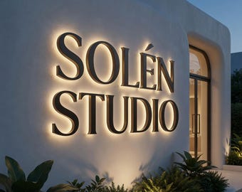Puede incluir: Una pared exterior blanca presenta las letras oscuras y tridimensionales SOLÉN STUDIO. Las letras están montadas con una luz de fondo que proyecta un brillo suave sobre la superficie texturizada. Hay plantas verdes en la base de la pared.
