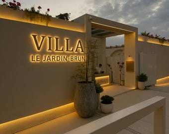 May include: A beige exterior wall features illuminated gold lettering that reads VILLA LE JARDIN-BRUN. The structure includes a recessed entrance, warm LED strip lighting, large grey stone planters with greenery, and a small reception desk.
