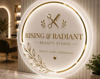 May include: A circular, backlit acrylic sign with a gold-toned frame. The sign features the text "RISING & RADIANT BEAUTY STUDIO" and "BEAUTY. CARE. CONFIDENCE." with a graphic of scissors, a comb, and leafy branches in gold lettering.