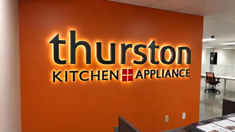 May include: A wall-mounted sign featuring the black text "thurston" in a large sans-serif font above the words "KITCHEN APPLIANCE" in a smaller font, with a red square icon between the words, all backlit against a vibrant orange wall.