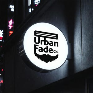 Puede incluir: Un letrero redondo e iluminado con el texto "Urban Fade co." en negro. El letrero presenta una navaja de barbero estilizada y un gr&aacute;fico de barba. Est&aacute; montado en una pared gris oscura, con luces borrosas de la ciudad en el fondo.