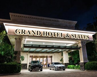 May include: The exterior entrance of the Grand Hotel & Suites at night, featuring a large illuminated sign with the text GRAND HOTEL & SUITES, supported by two fluted columns, with a black SUV and a silver sedan parked under the canopy.