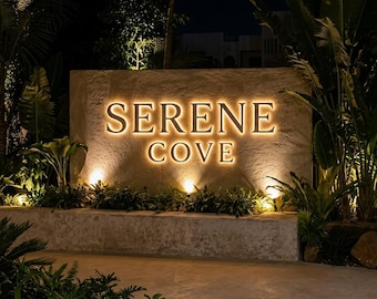 May include: A rectangular stone sign with the text SERENE COVE illuminated in warm white light. The sign is set against a textured wall, surrounded by lush green tropical plants and illuminated by small ground-level spotlights at night.