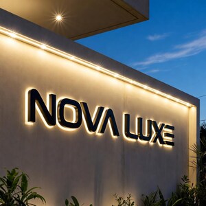 May include: A wall-mounted sign featuring the black text NOVA LUXE in a modern sans-serif font. The letters are backlit with a warm white glow, mounted on a smooth light gray concrete wall illuminated by a linear light fixture along the top edge.