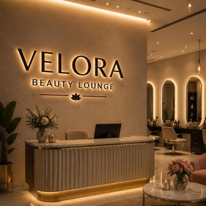 May include: A reception area for Velora Beauty Lounge featuring a cream-colored fluted desk with a marble top. On the wall, the black backlit sign reads VELORA BEAUTY LOUNGE with a lotus icon. The space includes oval mirrors, gold lighting, and plants.