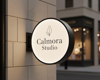 May include: A circular, wall-mounted, illuminated sign with a black frame. The white face features a black line-art leaf icon above the text "Calmora Studio" in a serif font. The sign is attached to a light-colored, tiled exterior wall surface.