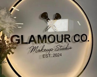 May include: A circular, illuminated sign featuring the black text GLAMOUR CO. Makeup Studio EST. 2024. Above the text is a graphic of two crossed makeup brushes. The sign has a thin, metallic gold-toned circular frame and a glowing light border.