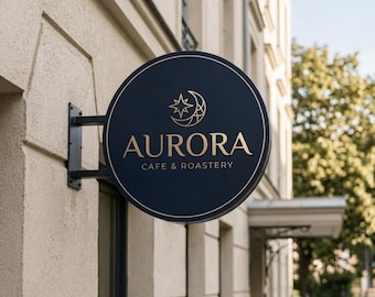 May include: A circular black metal sign mounted to a beige building wall. The sign features a gold-colored crescent moon and star icon above the text AURORA CAFE & ROASTERY in gold lettering, surrounded by a thin gold circular border line.