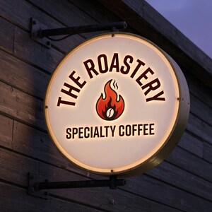 Puede incluir: Un letrero redondo e iluminado para "The Roastery Specialty Coffee" est&aacute; montado en una pared de madera. El letrero presenta un gr&aacute;fico de llama roja y naranja con un grano de caf&eacute;, y el texto est&aacute; en negrita, fuente serif.