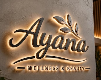May include: A wall-mounted sign featuring the black text Ayana in a large cursive font with a leaf graphic above the letter y. Below, the text WELLNESS & BEAUTY appears in a smaller sans-serif font. The sign is backlit with a warm glow.