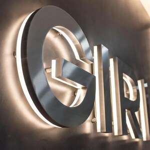 May include: A three-dimensional wall sign featuring the letters G, I, R, and I in a metallic silver finish. Each letter is mounted slightly away from the dark wall surface, with integrated white LED backlighting creating a glowing halo effect.