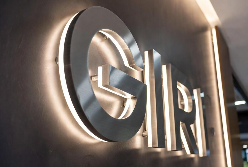 May include: A three-dimensional wall sign featuring the letters G, I, R, and I in a metallic silver finish. Each letter is mounted slightly away from the dark wall surface, with integrated white LED backlighting creating a glowing halo effect.