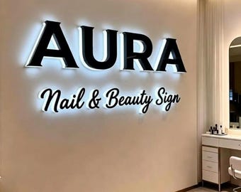 May include: A wall-mounted sign featuring the word AURA in large, bold, black, backlit capital letters, positioned above the words Nail & Beauty Sign in a smaller, black, cursive, backlit font, creating a glowing halo effect.