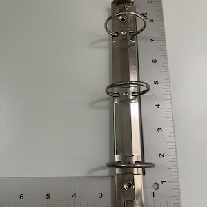 May include: A silver-colored binder mechanism with three metal rings. The binder is positioned vertically against a white background. A ruler is placed to the side, showing measurements in inches.
