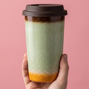 May include: A ceramic travel mug with a brown lid and a speckled green and orange body. The mug is held by a hand against a pink background. The mug is designed for hot or cold beverages.