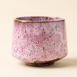 May include: A handcrafted ceramic cup with a textured, speckled surface. The cup features a gradient of colors, transitioning from a deep burgundy at the base to a lighter pink and white near the rim. The rim has a dark brown glaze.