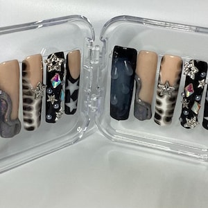 May include: A set of ten press-on nails displayed in a clear plastic case. The designs include black nails with raised droplets, nude nails with grey swirls, leopard print patterns, black nails with silver star charms, iridescent gems, and white stars.