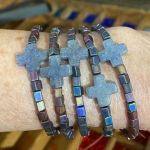 May include: A set of five beaded bracelets. Each bracelet features a light blue cross pendant and iridescent square beads. The bracelets are stacked on top of each other, showcasing the design and color variations.