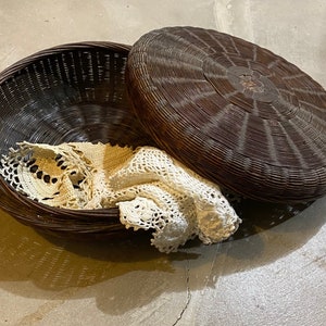 May include: A brown wicker basket with a lid, the lid is slightly ajar and a white crocheted doily is inside the basket.