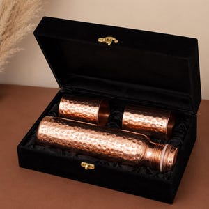 May include: A set consisting of one hammered copper water bottle and two matching hammered copper tumblers, all presented inside a black velvet-lined rectangular box with a gold-toned metal latch closure on the front edge.