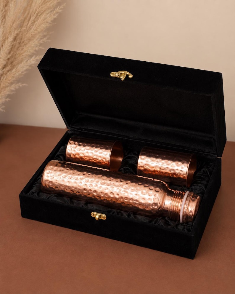 May include: A set consisting of one hammered copper water bottle and two matching hammered copper tumblers, all presented inside a black velvet-lined rectangular box with a gold-toned metal latch closure on the front edge.