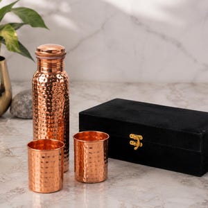May include: A hammered copper water bottle with a matching cap and two matching hammered copper tumblers. The set is accompanied by a rectangular black velvet storage box featuring a gold-colored metal latch on the front closure.