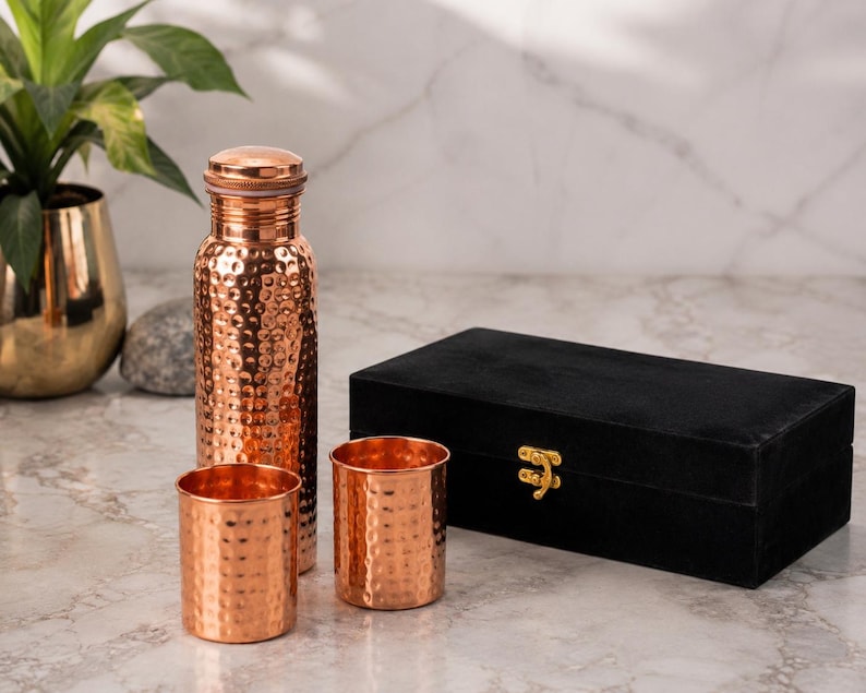 May include: A hammered copper water bottle with a matching cap and two matching hammered copper tumblers. The set is accompanied by a rectangular black velvet storage box featuring a gold-colored metal latch on the front closure.
